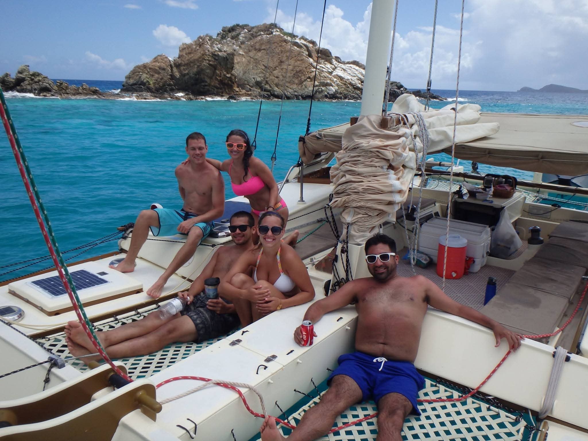 Happy group of five guests relaxing on the Mahiya catamaran deck with a rocky VI island and turquoise water behind