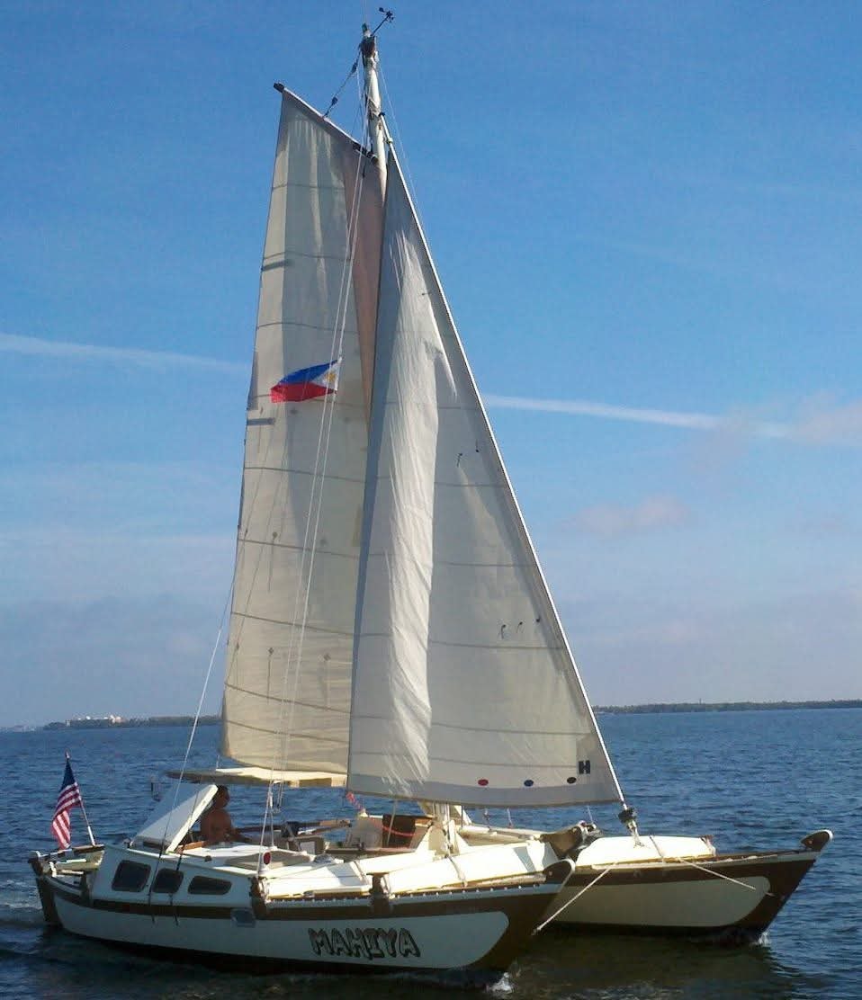 The Mahiya catamaran under full white sail with the Philippine flag flying, American flag at the stern, blue sky behind
