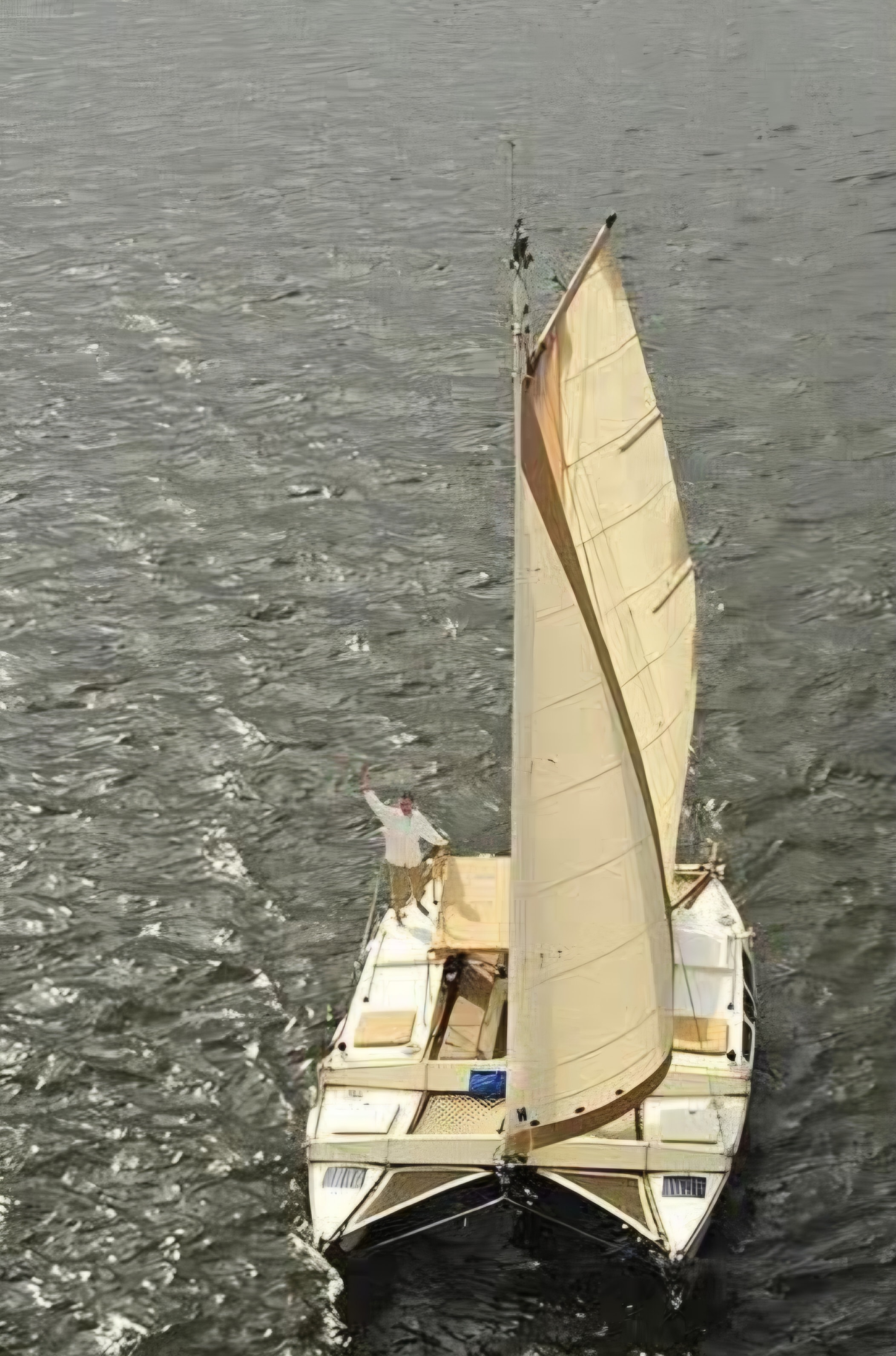 Aerial drone shot of the Mahiya catamaran under full cream sail on choppy grey water, person waving from the bow deck